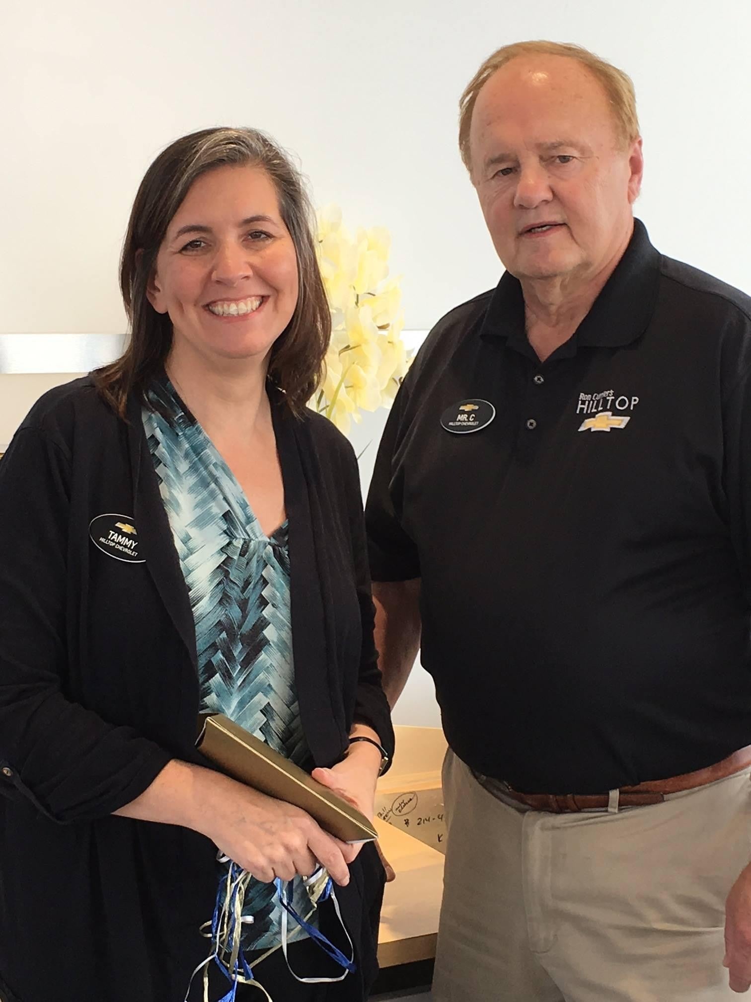 Two people standing side by side, one holding items, the other wearing a Hilltop polo shirt. Yellow flowers in the background.