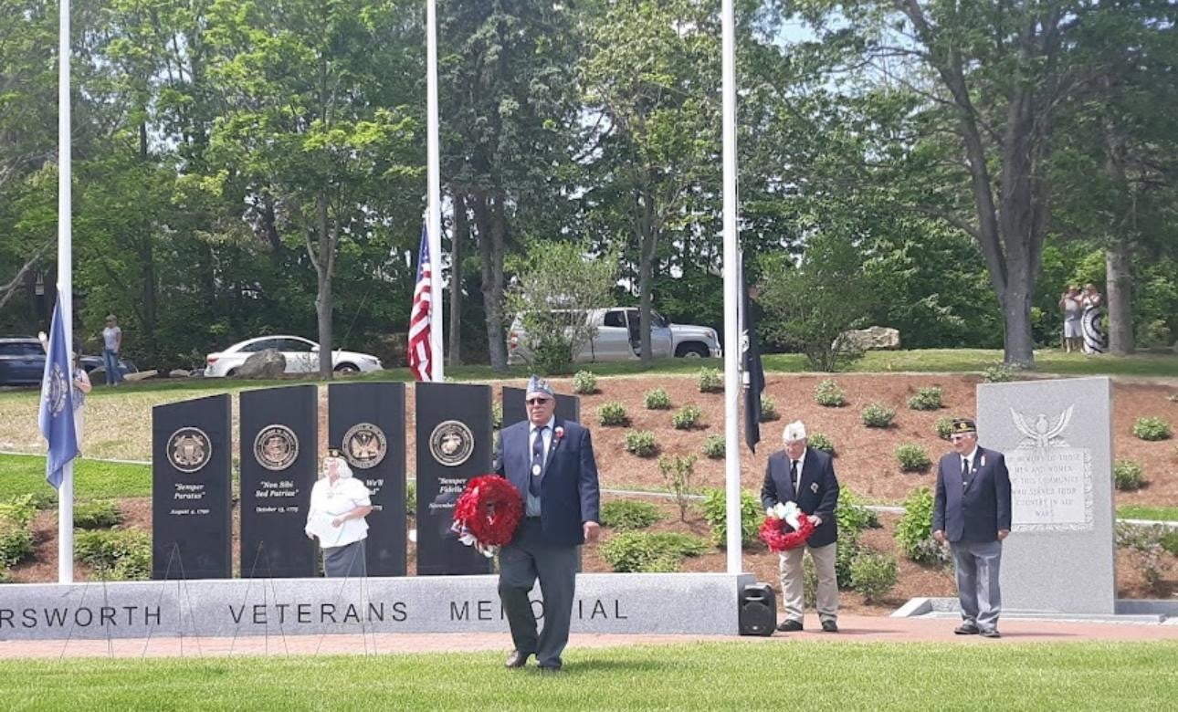 Group of people in front of a memorial with military emblems and inscriptions. Flags behind them.