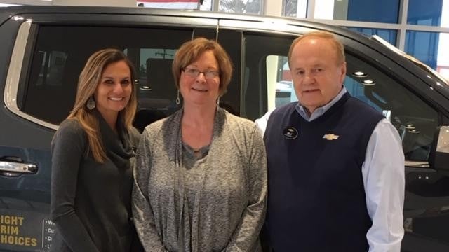 Three people stand in front of a Chevrolet vehicle inside a showroom. The person on the right is wearing a vest with the Chevrolet logo and name tag.