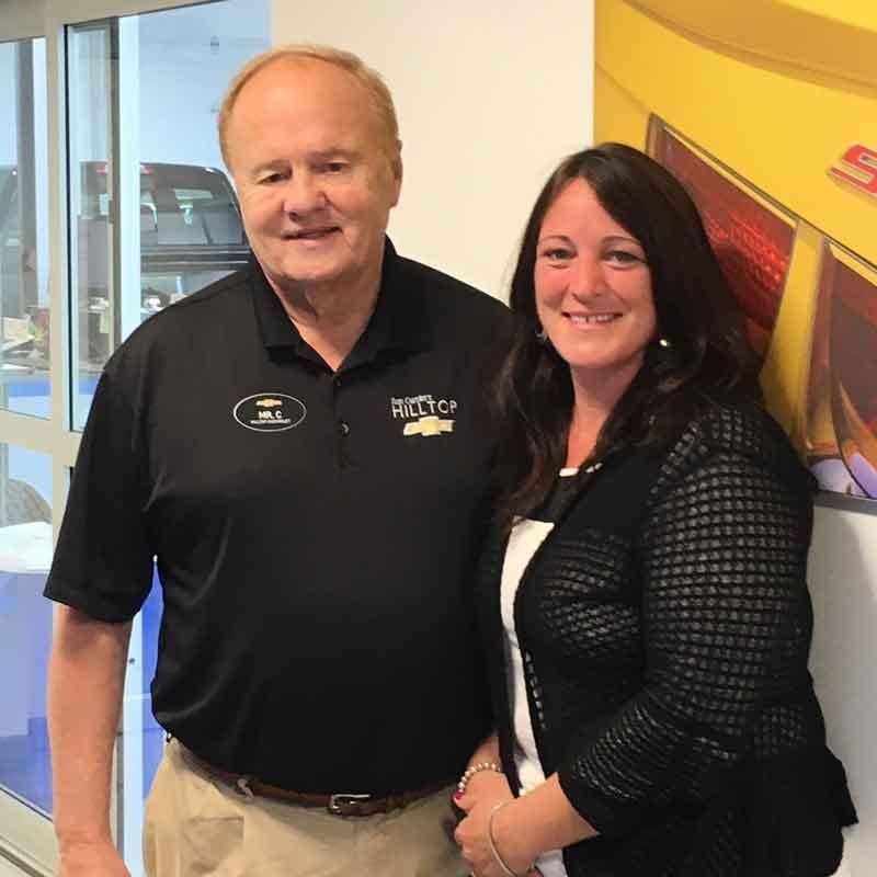 Two people stand together indoors. The person on the left is wearing a black polo shirt with a logo and name tag
