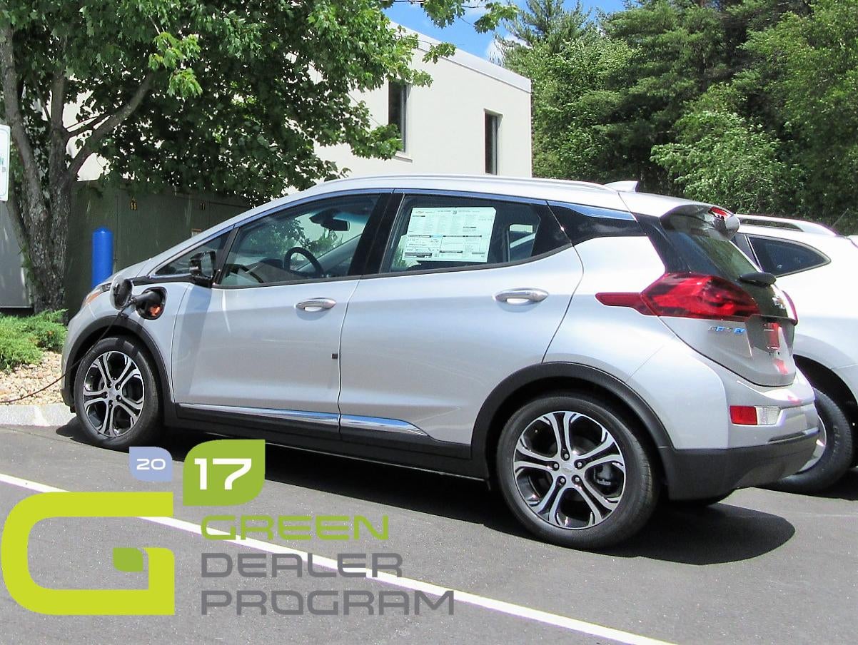 A silver electric car is parked outside with a charging cable plugged into its side. There is a 2017 Green Dealer Program logo in the bottom left corner.