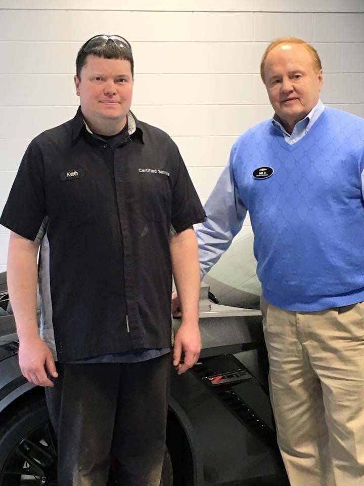 Two people standing in front of a car, one in a black shirt, the other in a blue sweater.